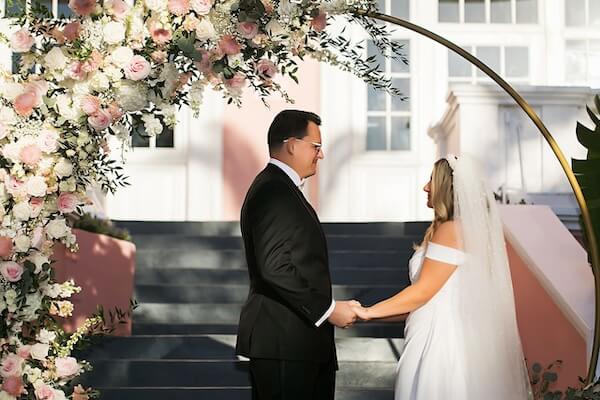 Bride and groom in their wedding attire holding hands in front of the moon gate arch wrapped in pink and white flowers during the wedding ceremony