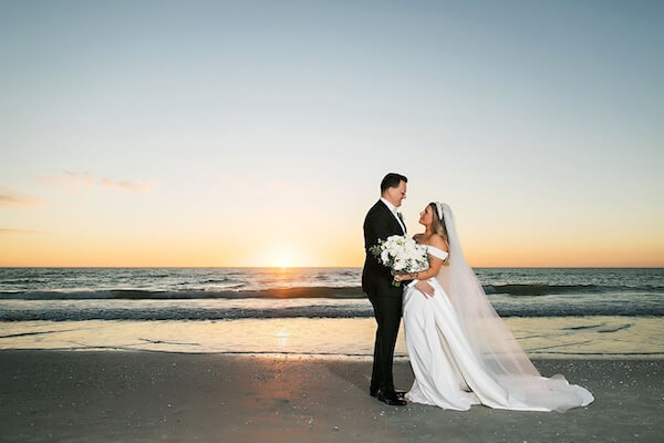 Panned out picture of the bride and groom on St. Pete beach at sunset, standing on the sand in front of the water gazing into each others eyes