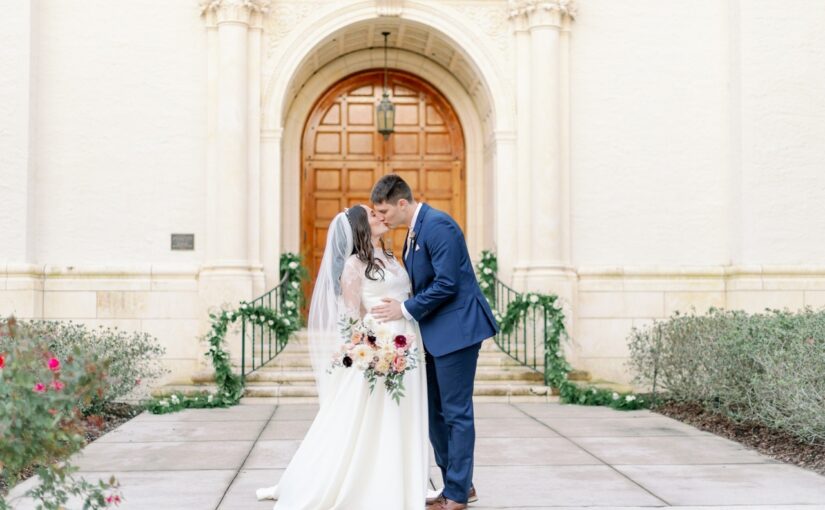 Bride and groom sharing a kiss outside the wedding venue while bride is also holding her bouquet in one hand