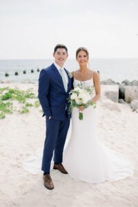 The bride and groom captured standing on the sand with the ocean in the background, dressed in their wedding attire and the bride holding her bouquet