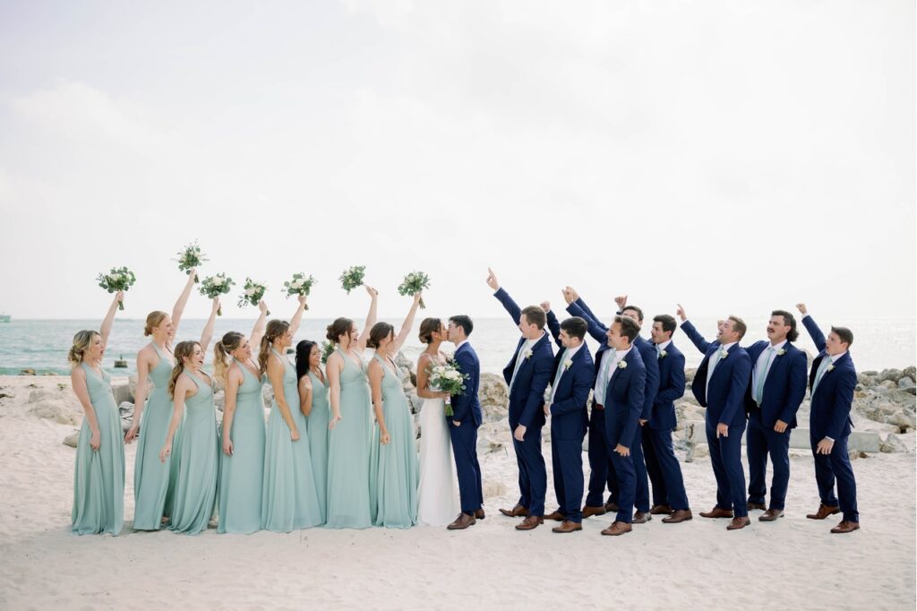 Entire wedding party on the beach dressed in their attire, cheering for the bride and groom as they share a kiss in front of the ocean