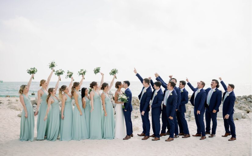 Entire wedding party on the beach dressed in their attire, cheering for the bride and groom as they share a kiss in front of the ocean