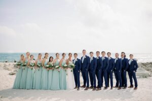 The entire wedding party in their wedding attire standing on the sand in front of the ocean taking a group picture