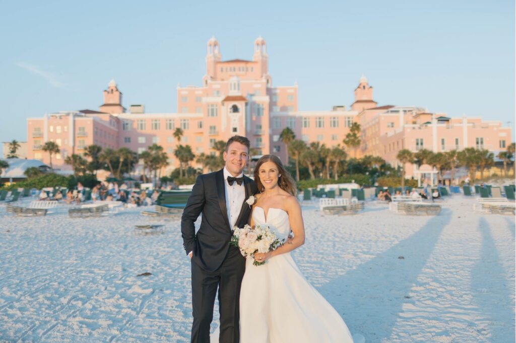 The bride in groom in wedding attire standing on the beach with the Don Cesar hotel in the background