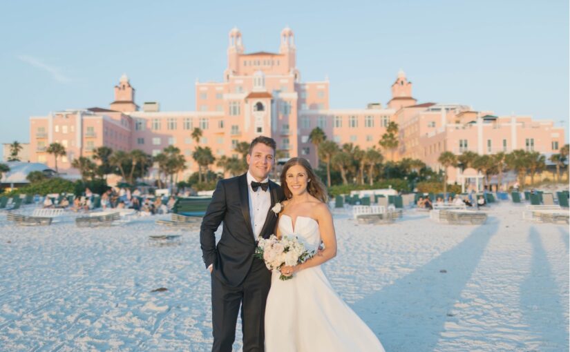 The bride in groom in wedding attire standing on the beach with the Don Cesar hotel in the background