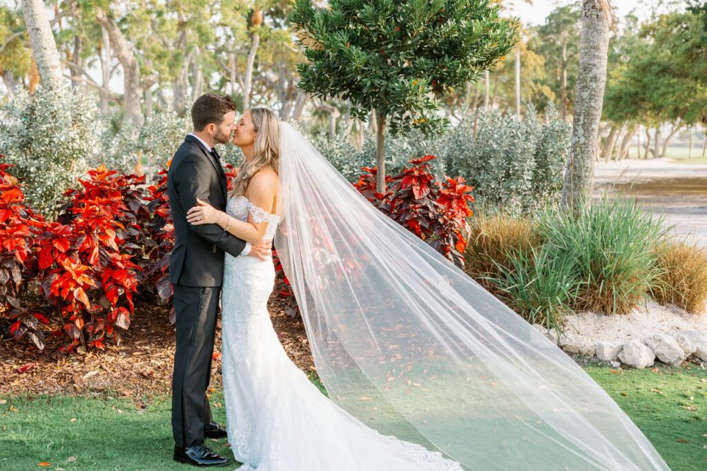 Bride and groom outdoors in front of red foliage sharing a kiss with the brides veil blowing in the wind