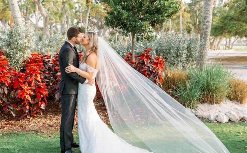 Bride and groom outdoors in front of red foliage sharing a kiss with the brides veil blowing in the wind