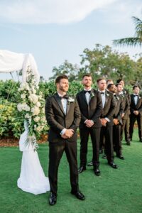 The groom waiting at the end of the aisle by the white wedding arch with his groomsmen behind him lined up