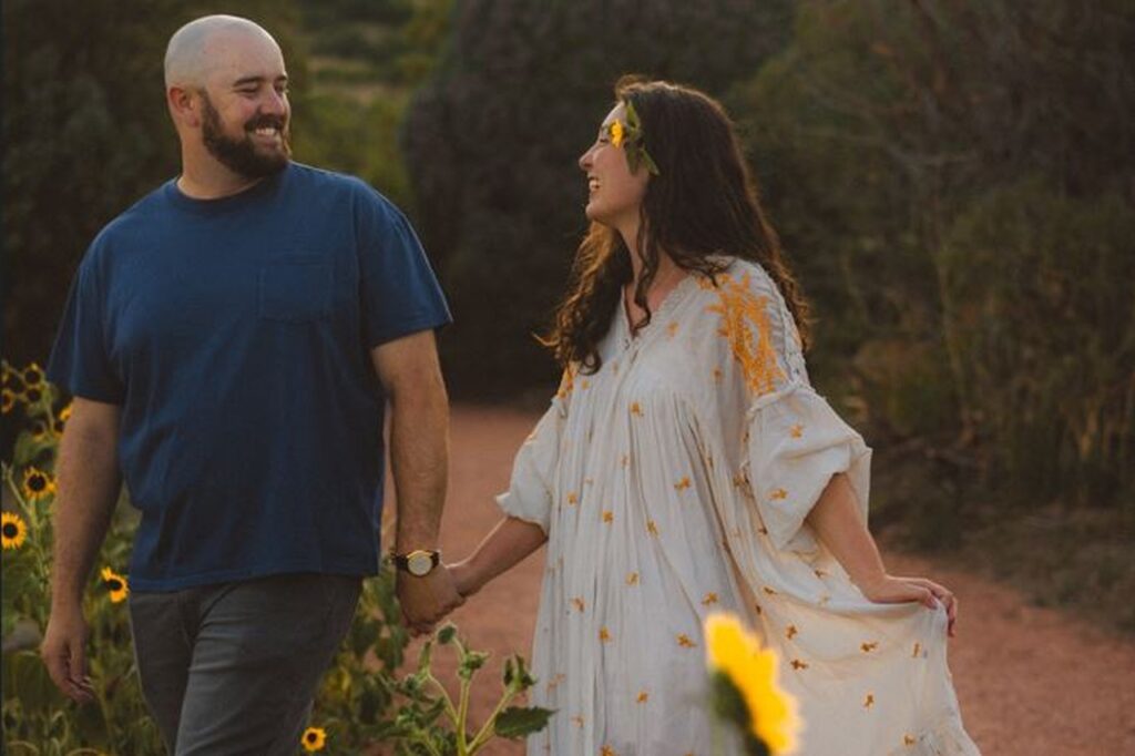 August Monthly Wedding winners holding hands in a field of sunflowers with the sunlight peaking through the trees in the background