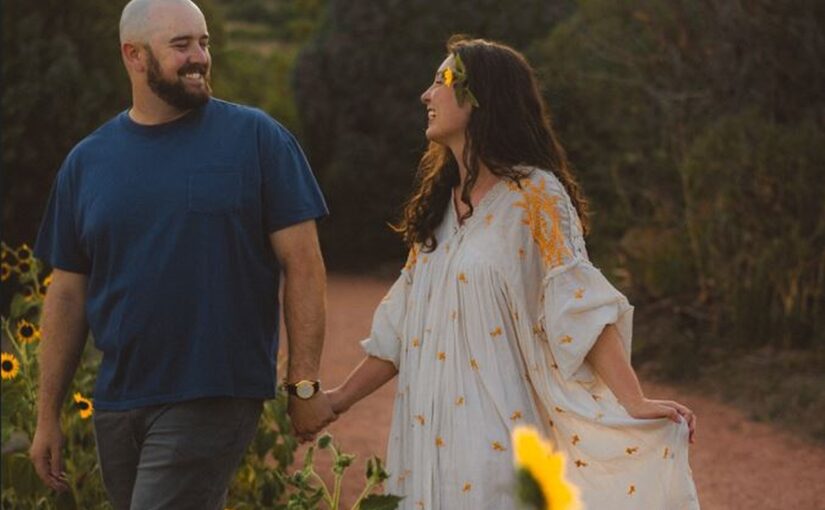 August Monthly Wedding winners holding hands in a field of sunflowers with the sunlight peaking through the trees in the background