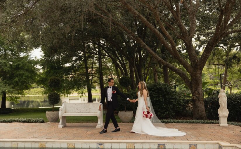 Bride and Groom hand in hand outdoors in their wedding attire taking a photo in front of a giant tree and a pond in the background