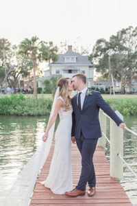 The bride and groom in their wedding attire sharing a kiss on the dock with the house in the background