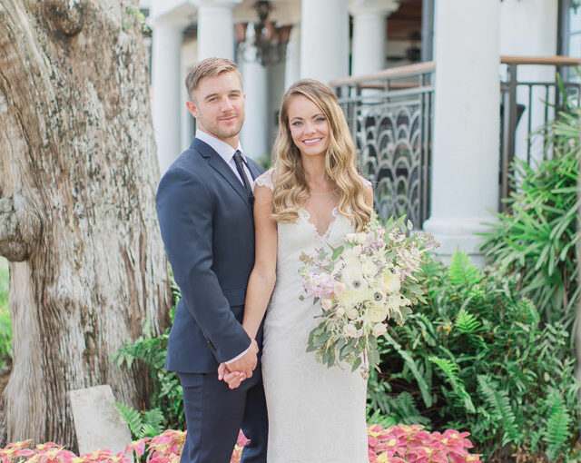 The bride and groom in their wedding attire standing next to a tree while holding hands and posing for a picutre. The bride is holding her bouquet in the opposite hand