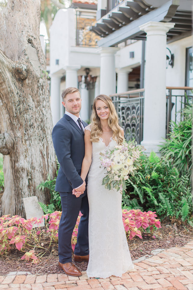 The bride and groom in their wedding attire standing next to a tree while holding hands and posing for a picutre. The bride is holding her bouquet in the opposite hand