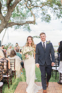 The bride and groom arm in arm walking out of the wedding ceremony with their guests still seated in their chairs and a tree and water in the background of the photo