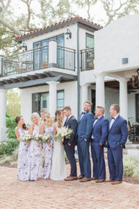 The bride and groom in the center of all their wedding party gazing at each other as they stand in a line on the cobblestone driveway while all dressed in their wedding attire