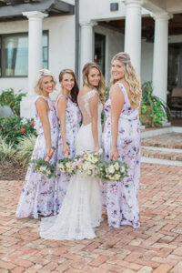 The bride in her wedding gown and the bridesmaids in their dresses all holding their bouquets while standing in a line looking over their shoulders as they pose for a picture while standing on the cobblestone driveway