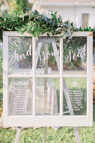 Find your seat written on a window with greenery on top of it and all the guests names and seating arrangement written on it