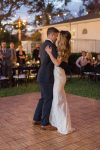 The bride and groom on the dancefloor having their first dance as the sun is setting and the guests watch as they sway under the string lights on the wooden dance floor