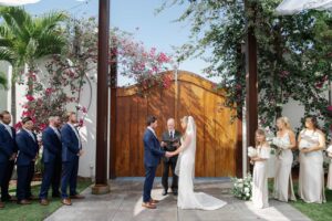 The bride and groom holding hands in front of a wooden door, with their wedding party on either side of them as they stand outdoors at the wedding ceremony