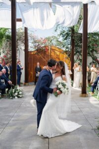 The groom kissing his bride as they exit the wedding ceremony as guests are all around and they are standing under billowing white linens hanging from steel beams