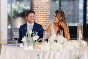 The bride and groom in their wedding attire sitting at their table draped in white linen with flowers