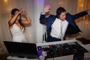 The bride and groom in wedding attire at their reception behind the DJ booth dancing and pretending to DJ