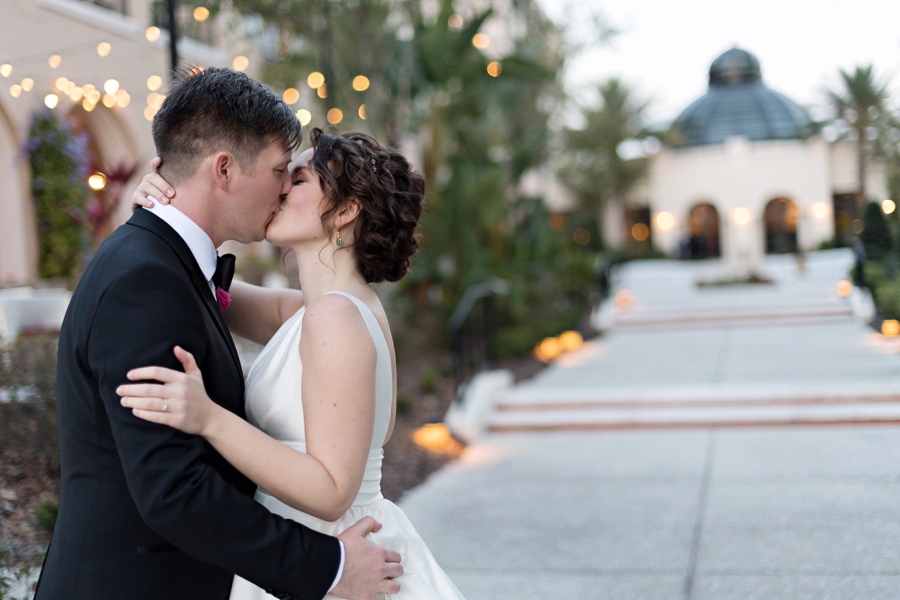 The bride and groom pictured to the left sharing a kiss outside the venue, with twinkling lights in the background of the photo.