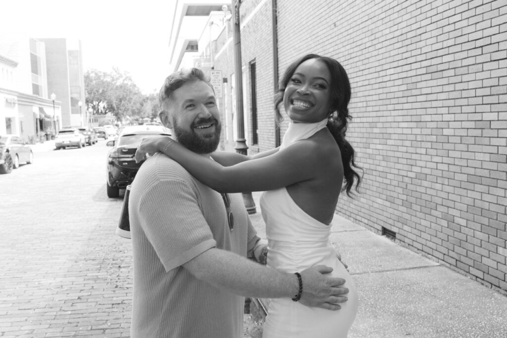 A black and white photo of Decembers Monthly Wedding Winners, with the girls arms around the guys shoulders and his hands around her hips. They pose for a picture on the sidewalk