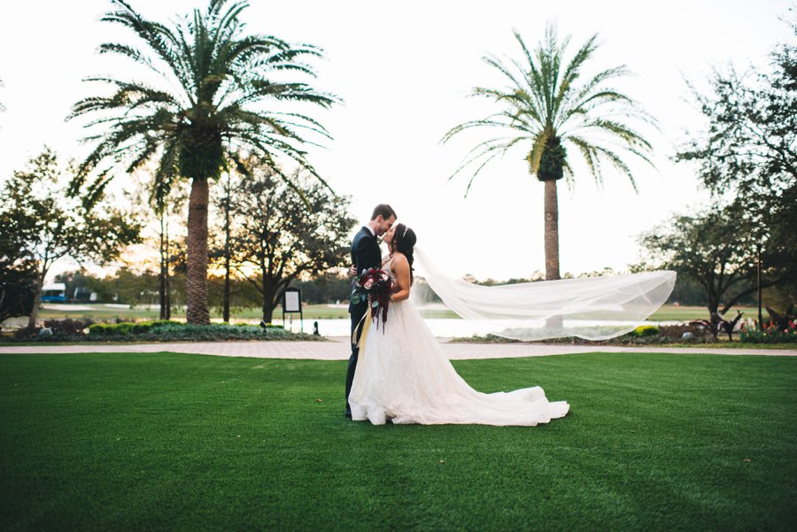 The bride and groom standing in the grass both dressed in wedding attire sharing a kiss with the brides veil blowing in the wind behind her