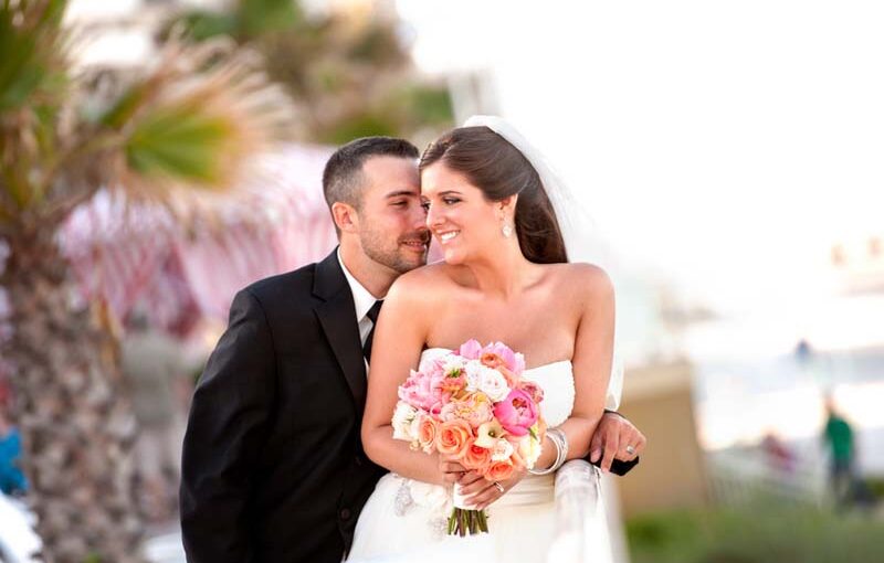 The groom standing outside behind the bride as they bother wear their wedding attire. The bride turning her head into his as she is holding her bouquet, with the blurred background of outside