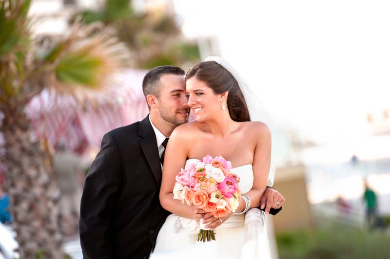 The groom standing outside behind the bride as they bother wear their wedding attire. The bride turning her head into his as she is holding her bouquet, with the blurred background of outside