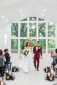 The second bride and groom holding hands and walking back down the aisle as if they just finished their wedding ceremony, as the vendors are sitting in the chairs watching.