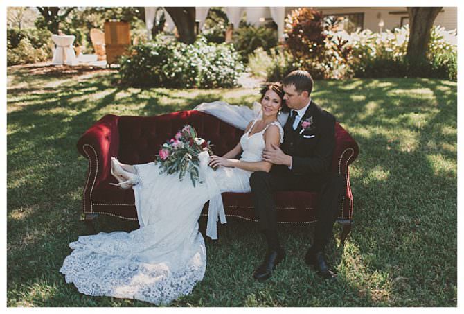 The bride and groom in wedding attire sitting outside on a red velvet couch, with the bride laying across it onto the grooms lap