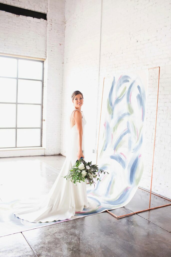 Bride looking over her shoulder as she poses for camera, while standing in front of aisle runner and holding her bouquet down to the side of her