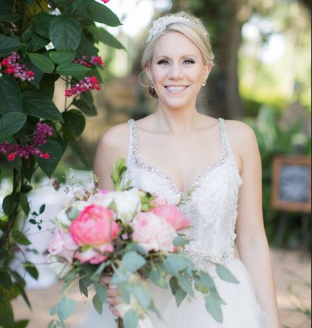 Bride wearing a wedding gown while holding bouquet of pink white and greenery. While standing next to the outdoor greenery