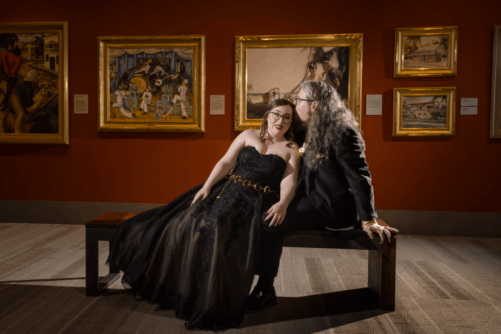 The bride and groom in their black wedding attire sitting on a bench as the bride lays across it onto the grooms lap with the portraits in the museum in the background