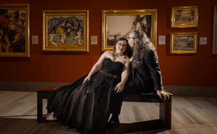 The bride and groom in their black wedding attire sitting on a bench as the bride lays across it onto the grooms lap with the portraits in the museum in the background