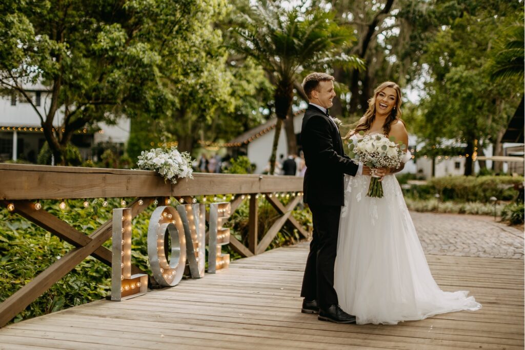 The bride in groom in their wedding attire standing outside the venue at the Cross Creek Ranch on a wooden bridge in each others arms with a Love sign off to the left of them as they smile for a photo