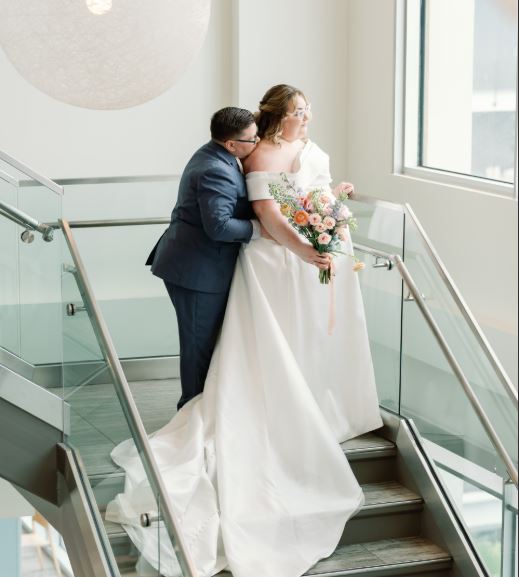 February Monthly Wedding Winner dressed in wedding attire with the bride in front and the groom standing behind the bride with his head resting near her shoulder while they stand on stairs and gaze out the window