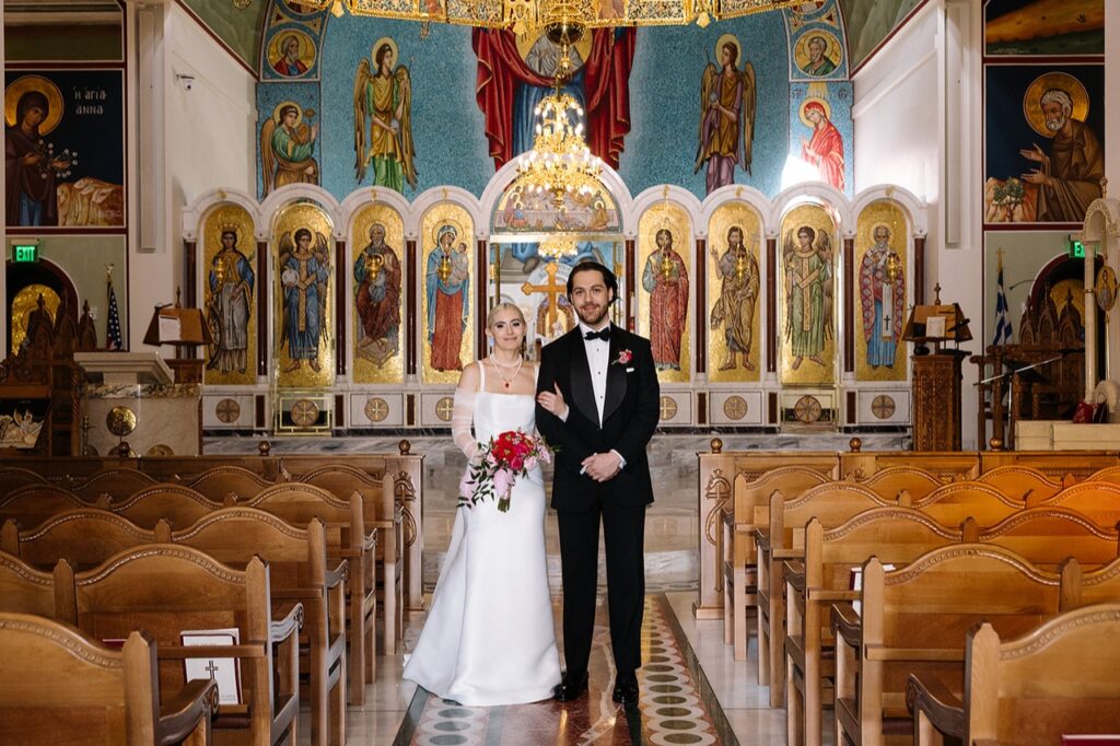 Kali and Zach Doyle on their wedding day May 25, 2024. Dressed in their wedding attire standing in the aisle at the Holy Trinity Orthodox Church