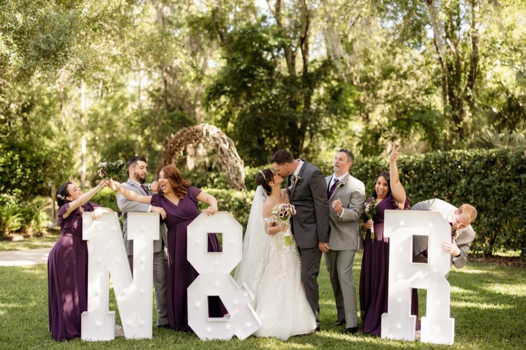 Noelis and Ryan wedding at Bakers Ranch 4/7/24. Bride and groom sharing a kiss in front of large letters with people from the wedding standing next to them posing for the outdoor photo